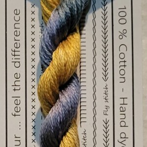 Pode incluir: Um novelo de fio de bordado de algodão trançado em tons de azul e amarelo. O fio é etiquetado como "VD 14 Billy Button" e é 100% algodão, tingido à mão na Austrália. A etiqueta também indica que o fio tem 10 metros de comprimento e é pré-cortado em comprimentos de 50 cm.