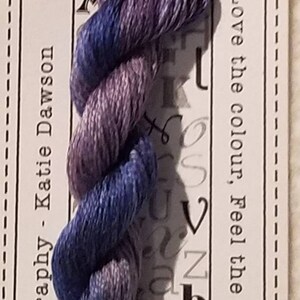 May include: A skein of hand-dyed embroidery floss in shades of blue and purple. The floss is labeled "Charlie" and is 10 meters long. The label also says "Stranded Cotton" and "Pre-Cut".