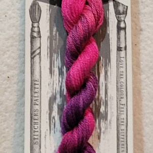 May include: A skein of hand-dyed embroidery floss in shades of pink and purple. The floss is labeled "Zinnia" and is part of the "Stitchers Palette" collection. The label also says "10m E-Cut" and "Love the Colour, Feel the Difference."