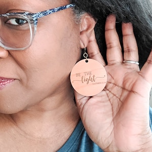 May include: Wooden earrings with the text "Be the light" and "Matthew 5:14" in a cursive font. The earrings are a light brown color and have a round shape.