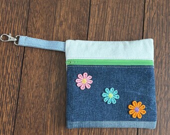 May include: A denim pouch with a light blue top and a green zipper. The pouch features three embroidered flower appliques in pink, blue, and orange. A light blue strap with a silver clasp is attached.