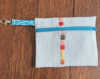 May include: A light blue denim pouch with a turquoise zipper and a matching wrist strap. The pouch features a vertical embroidered design in various colors. The wrist strap is patterned with white dots.