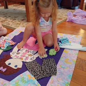 Op de afbeelding: Een jong meisje speelt met een poppenquilt. De quilt is gemaakt van verschillende stoffen en heeft een paarse achtergrond. De pop is gemaakt van vilt en heeft bruin haar, bruine ogen en een rode mond. Het meisje draagt een roze shirt en een blauwe broek. Ze houdt een bloem in haar hand.