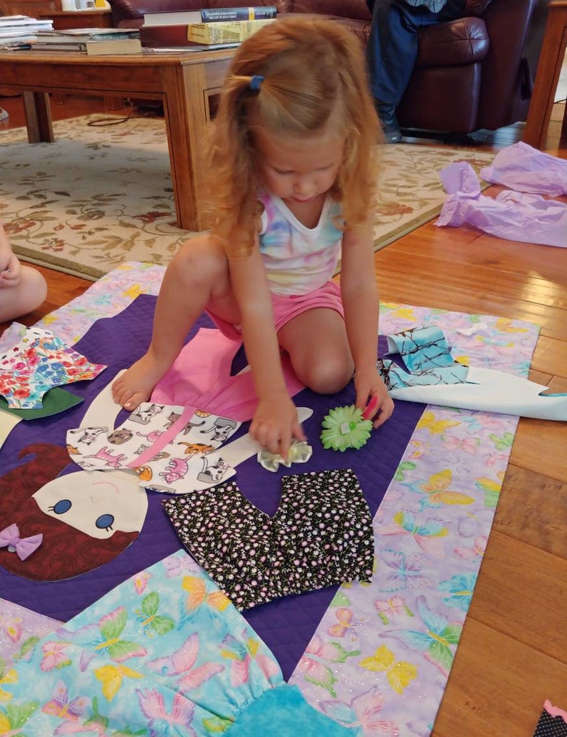 Op de afbeelding: Een jong meisje speelt met een poppenquilt. De quilt is gemaakt van verschillende stoffen en heeft een paarse achtergrond. De pop is gemaakt van vilt en heeft bruin haar, bruine ogen en een rode mond. Het meisje draagt een roze shirt en een blauwe broek. Ze houdt een bloem in haar hand.