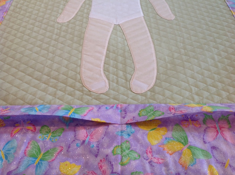 Op de afbeelding: Een lichtgroen gestikt babydekentje met een roze vilt poppenapplicatie. Het dekentje heeft een paarse en witte rand met een vlinderprint.