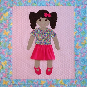 Op de afbeelding: Een handgemaakte, gestikte wandhanger met een poppenfiguur van een meisje met bruin haar, een roze strik, een shirt met bloemenprint, een roze rok en bruine schoenen. De pop is omringd door een blauwe en witte vlinderprint rand.