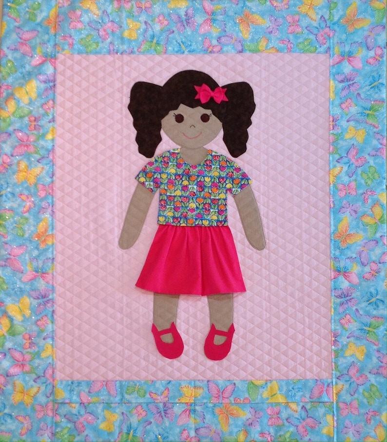 Op de afbeelding: Een handgemaakte, gestikte wandhanger met een poppenfiguur van een meisje met bruin haar, een roze strik, een shirt met bloemenprint, een roze rok en bruine schoenen. De pop is omringd door een blauwe en witte vlinderprint rand.