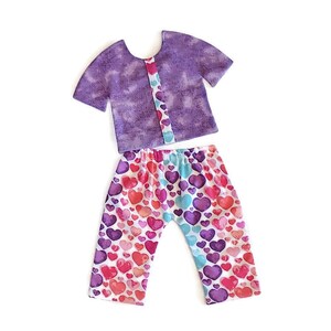 May include: A purple short-sleeved shirt with a white heart pattern on the front and a pair of pants with a white background and a colorful heart pattern.