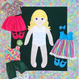 May include: A felt doll with a white dress and blonde hair. The doll is surrounded by felt clothing and accessories, including a pink skirt, a blue and white striped dress, a green pair of pants, and a plaid shirt. The doll is on a green background with a butterfly pattern.
