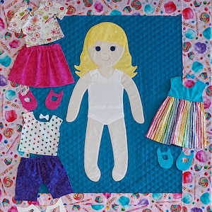 May include: A blue quilted fabric with a white doll in the center. The doll has blonde hair, a white dress, and is wearing different outfits. The fabric is surrounded by a pink border with candy and lollipop designs.