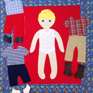 May include: A felt doll with blonde hair and a white undershirt, along with a variety of felt clothing items, including a red shirt, blue and white striped shirt, plaid shirt, red pants, blue pants, khaki pants, and brown boots. The doll and clothing are arranged on a red felt background with a blue and white patterned border.