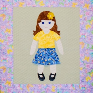 Op de afbeelding: Een handgemaakte, gestikte wandhanger met een viltpop met bruin haar, een geel shirt, een blauw-witte bloemenrok en zwarte schoenen. De pop is omgeven door een paarse rand met een vlinderpatroon.