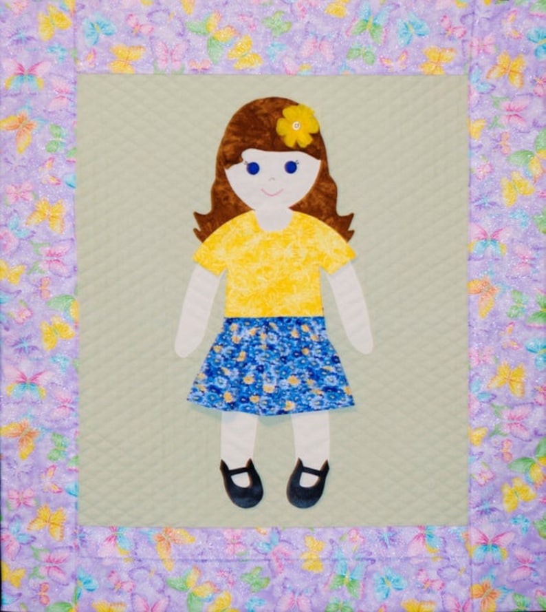 Op de afbeelding: Een handgemaakte, gestikte wandhanger met een viltpop met bruin haar, een geel shirt, een blauw-witte bloemenrok en zwarte schoenen. De pop is omgeven door een paarse rand met een vlinderpatroon.