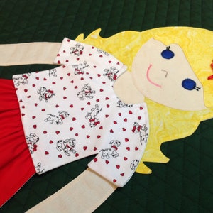 Op de afbeelding: Een handgemaakte pop met blond haar, een rode rok en een wit shirt met een patroon van rode hartjes en zwart-witte dalmati&euml;rpups. De pop heeft een geschilderd gezicht met blauwe ogen en een glimlach.