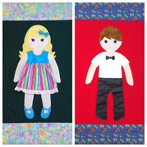May include: Two felt dolls, one with blonde hair wearing a pink and blue dress and the other with brown hair wearing a white shirt and black pants. The dolls are on a red and green background with a patterned border.