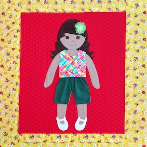 Op de afbeelding: Een handgemaakte vilten pop met bruin haar, een groen en roze geruit shirt, groene shorts en een bloem in haar haar. De pop staat op een rode achtergrond met een gele en witte rand.