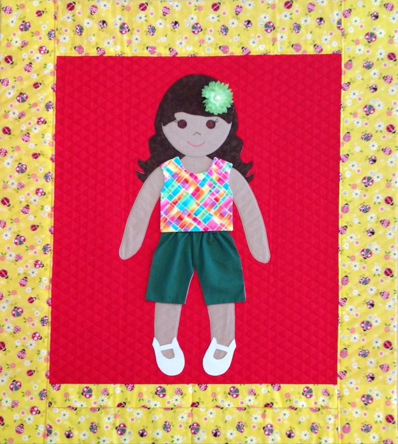 Op de afbeelding: Een handgemaakte vilten pop met bruin haar, een groen en roze geruit shirt, groene shorts en een bloem in haar haar. De pop staat op een rode achtergrond met een gele en witte rand.