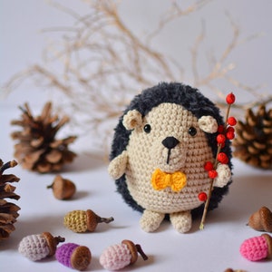 May include: A crocheted hedgehog toy wearing a yellow bow tie and holding a twig with red berries. The hedgehog is surrounded by acorns and pine cones.