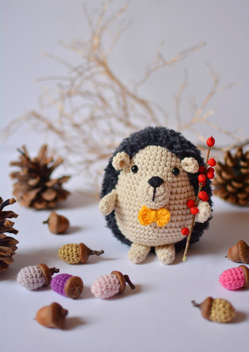 May include: A crocheted hedgehog toy wearing a yellow bow tie and holding a twig with red berries. The hedgehog is surrounded by acorns and pine cones.