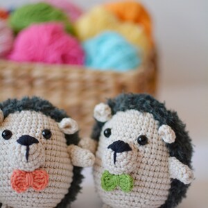May include: Two crocheted hedgehog plushies with black eyes and noses. One has a green bow tie and the other has an orange bow tie. The plushies are sitting on a white surface.