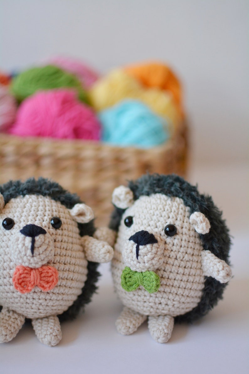 May include: Two crocheted hedgehog plushies with black eyes and noses. One has a green bow tie and the other has an orange bow tie. The plushies are sitting on a white surface.