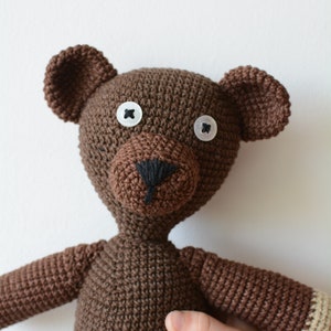May include: A brown crocheted teddy bear with white button eyes and a black stitched nose. The bear is standing on a white surface.
