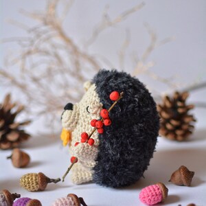 May include: A crocheted hedgehog toy holding a twig with red berries. The hedgehog is black and white with a yellow nose. The toy is surrounded by small brown acorns.