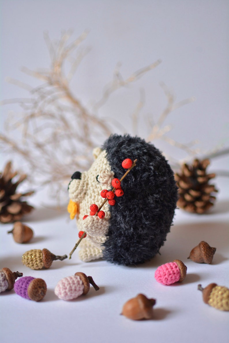 May include: A crocheted hedgehog toy holding a twig with red berries. The hedgehog is black and white with a yellow nose. The toy is surrounded by small brown acorns.