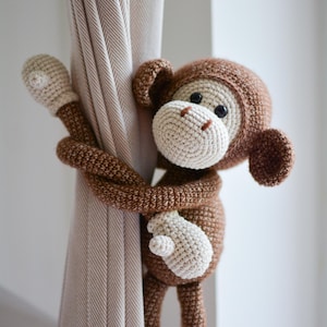 Puede incluir: Un amarre de cortina de mono de ganchillo en marrón y crema. El mono tiene una cara, orejas y cabeza de color marrón claro, con ojos oscuros y un hocico y manos de color crema. El mono está envuelto alrededor de una cortina beige.