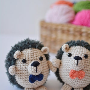 May include: Two crocheted hedgehog toys with black eyes and noses. One hedgehog is wearing a blue bow tie and the other is wearing an orange bow tie.