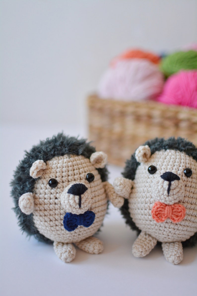 May include: Two crocheted hedgehog toys with black eyes and noses. One hedgehog is wearing a blue bow tie and the other is wearing an orange bow tie.