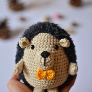 May include: A crocheted hedgehog plush toy with a black and white face and a yellow bow tie. The toy is being held in a person's hand.