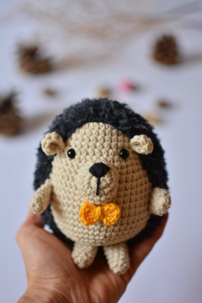 May include: A crocheted hedgehog plush toy with a black and white face and a yellow bow tie. The toy is being held in a person's hand.