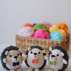 May include: Three crocheted hedgehog toys with different colored bow ties. The hedgehogs are sitting on a white surface in front of a basket of colorful yarn.