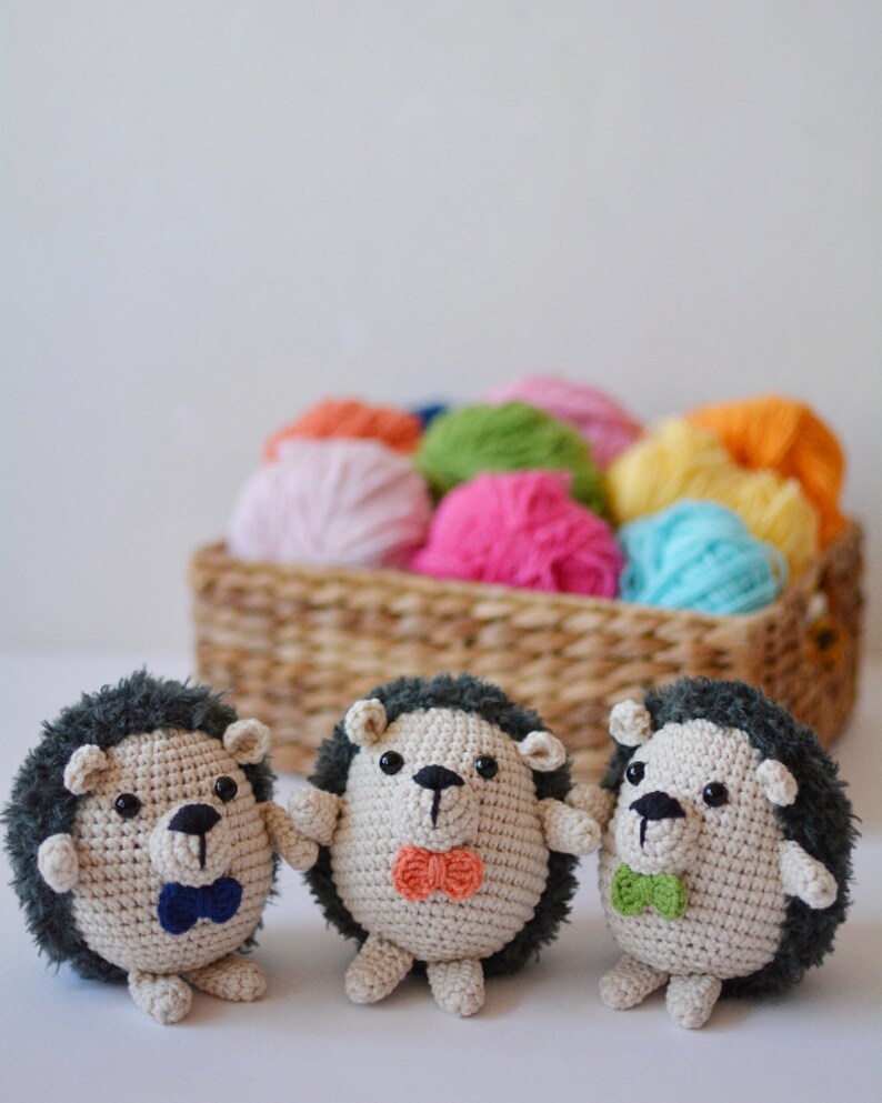 May include: Three crocheted hedgehog toys with different colored bow ties. The hedgehogs are sitting on a white surface in front of a basket of colorful yarn.