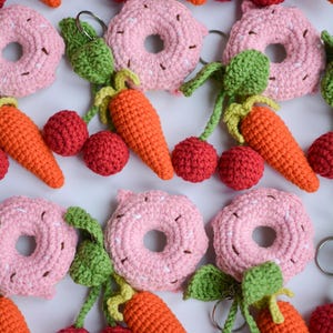Könnte beinhalten: Eine Sammlung handgefertigter Häkel-Schlüsselanhänger mit verschiedenen Lebensmitteln. Die Schlüsselanhänger umfassen rosa Donuts mit weißen Streuseln, orangefarbene Karotten und rote Kirschen mit grünen Stielen und Blättern. Jeder Schlüsselanhänger hat einen silbernen Ring.