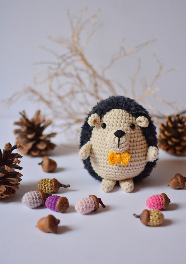 May include: A crocheted hedgehog toy with a yellow bow tie, surrounded by small crocheted acorns in various colors. The hedgehog is beige with a black and gray spiky back.