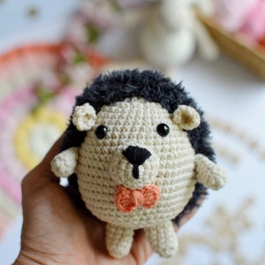 May include: A crocheted hedgehog toy with black fur, brown eyes, and a pink bow tie. The toy is being held in a hand.