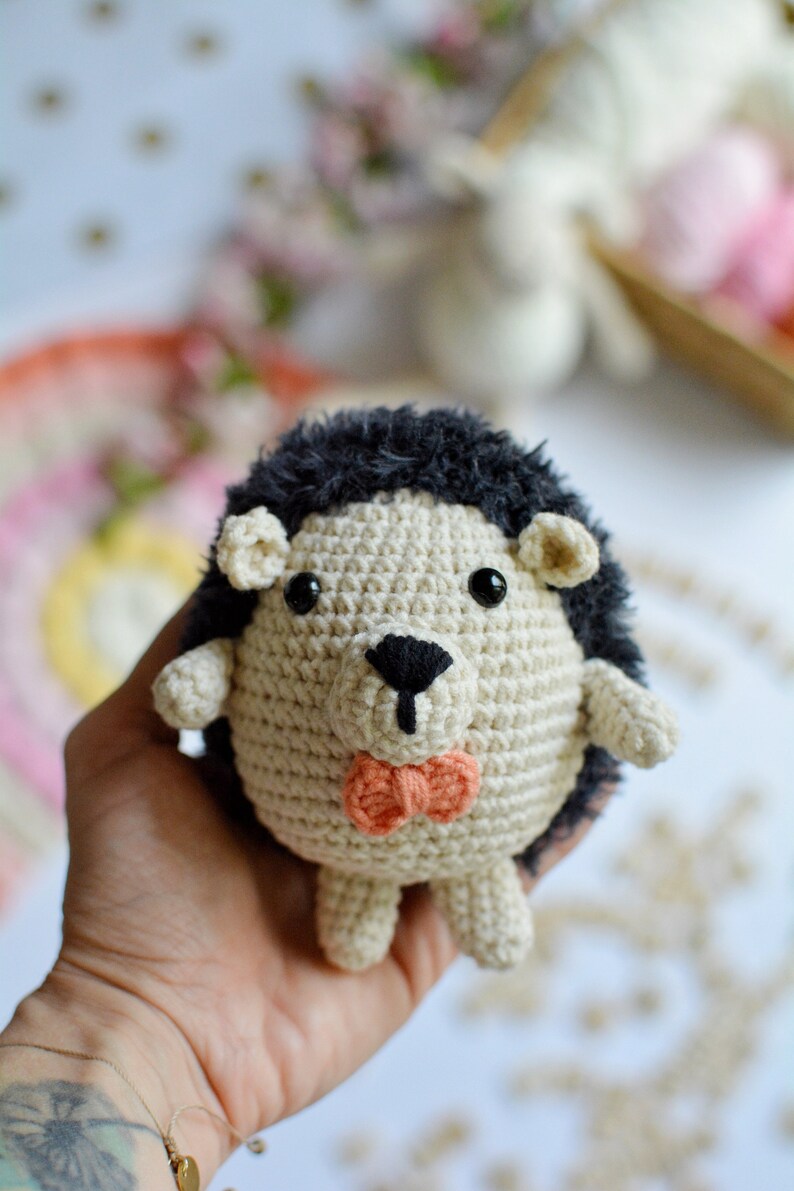May include: A crocheted hedgehog toy with black fur, brown eyes, and a pink bow tie. The toy is being held in a hand.