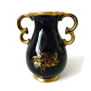 Può includere: Un vaso in ceramica nera con dettagli dorati e un motivo floreale. Il vaso ha due manici e un corpo arrotondato.