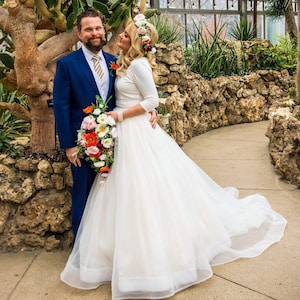 Puede incluir: Una novia y un novio posan para una foto de boda en un invernadero. La novia lleva un vestido de novia blanco con una corona de flores y el novio lleva un traje azul con una corbata dorada. Están de pie frente a una gran planta de cactus.