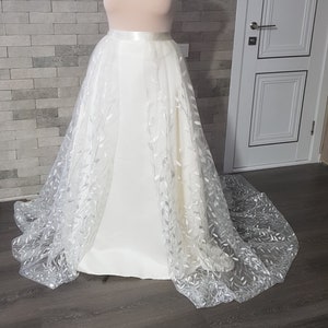 Puede incluir: Una falda de novia blanca con una superposición de delicado patrón de hojas. La falda está hecha de tul y tiene una cintura de satén.