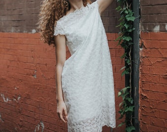 May include: A young woman with curly hair wearing a white lace dress stands in front of a brick wall with green ivy growing up it. She is looking up and reaching towards the ivy.