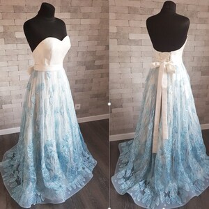 May include: Two light blue lace gowns displayed on mannequins. One gown is strapless with a sweetheart neckline and a white bodice. The other gown is backless with a white ribbon sash. Both gowns have a light blue lace overlay and a white underlay. The gowns are shown against a textured white brick wall.