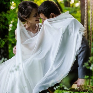 May include: A bride and groom playfully hide under a sheer white veil on a rustic rope swing. The couple is positioned in a lush green forest setting, surrounded by natural foliage. The veil drapes over them, creating a romantic and whimsical scene. The overall theme is wedding photography, capturing a candid moment of joy and intimacy between the newlyweds.