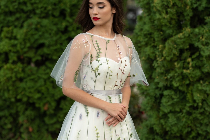 May include: A white tulle dress with floral embroidery and a sheer overlay. The dress has a fitted bodice and a full skirt. The model is wearing a light blue ribbon around the waist.