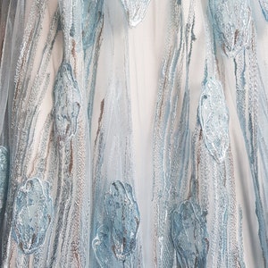 May include: Close-up view of a light blue tulle fabric with embroidered floral details. The embroidery features light blue and silver-toned floral motifs on a sheer, light blue background. The fabric has a soft, flowing texture, and the embroidery adds a delicate, elegant touch.  The design is reminiscent of a dress or skirt fabric.