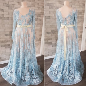 May include: Two light blue embroidered tulle maxi dresses with long sleeves and a fitted waistline. One dress is shown from the front and the other from the back. Both dresses feature a delicate floral pattern and a light yellow sash. The dresses are displayed on mannequins against a textured stone wall background.