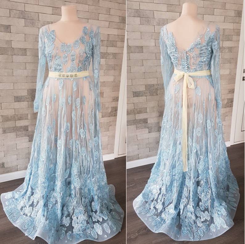 May include: Two light blue embroidered tulle maxi dresses with long sleeves and a fitted waistline. One dress is shown from the front and the other from the back. Both dresses feature a delicate floral pattern and a light yellow sash. The dresses are displayed on mannequins against a textured stone wall background.