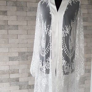 May include: White lace bridal robe with a satin lining. The robe has long sleeves and a relaxed fit.  Delicate floral lace patterns adorn the sheer fabric. The robe is displayed on a mannequin against a textured white brick wall. This elegant, lightweight garment is perfect for bridal preparations or special occasions.
